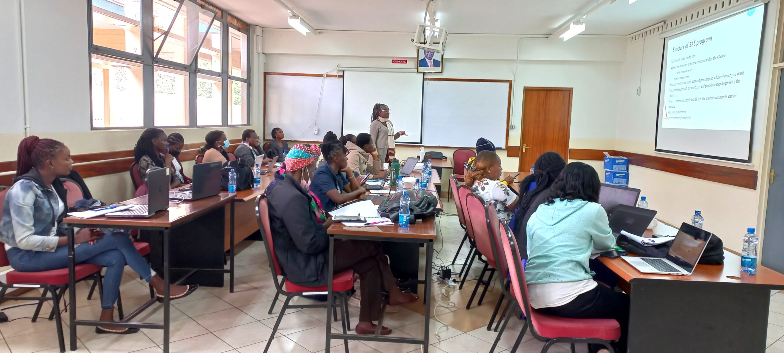 A classroom full of African students sit in rows facing a projector screen while an African female scholar lectures