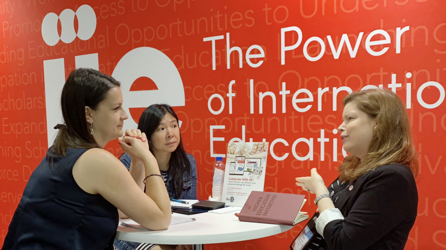 3 women meeting at table at IIE NAFSA booth with red banner in background