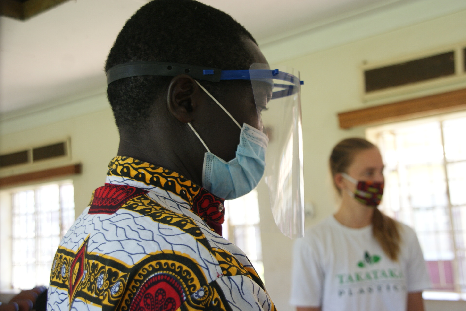 Man wears face shield and mask in foreground, Paige Balcom in background