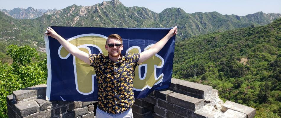 Male student stands holding University of Pittsburg flag on a rooftop with mountains in the background.