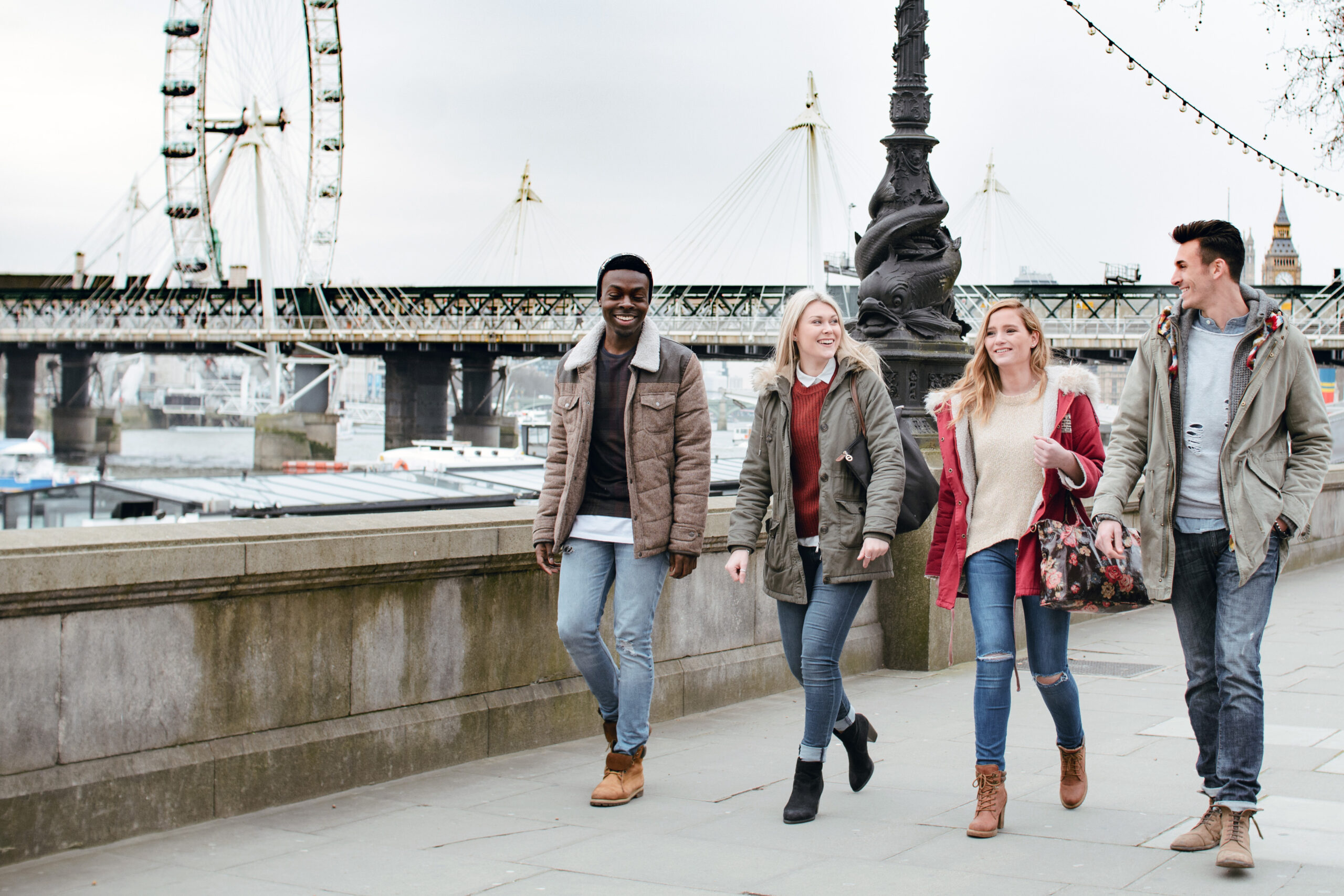 Group of four students walking alongside water and boats with a bridge in the background