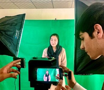 A woman stands in front of a green screen while two others record on a video camera